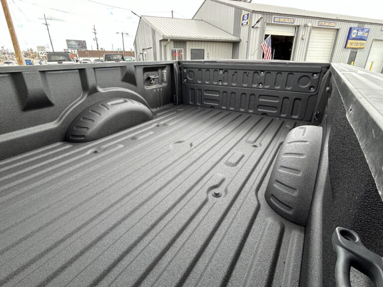 View of a truck bed that has been professionally coated with a mil-spec spray-on bedliner for enhanced protection. The bedliner appears black and textured. In the background is a shop advertising "4x4 Specialists" and "TNT Customs