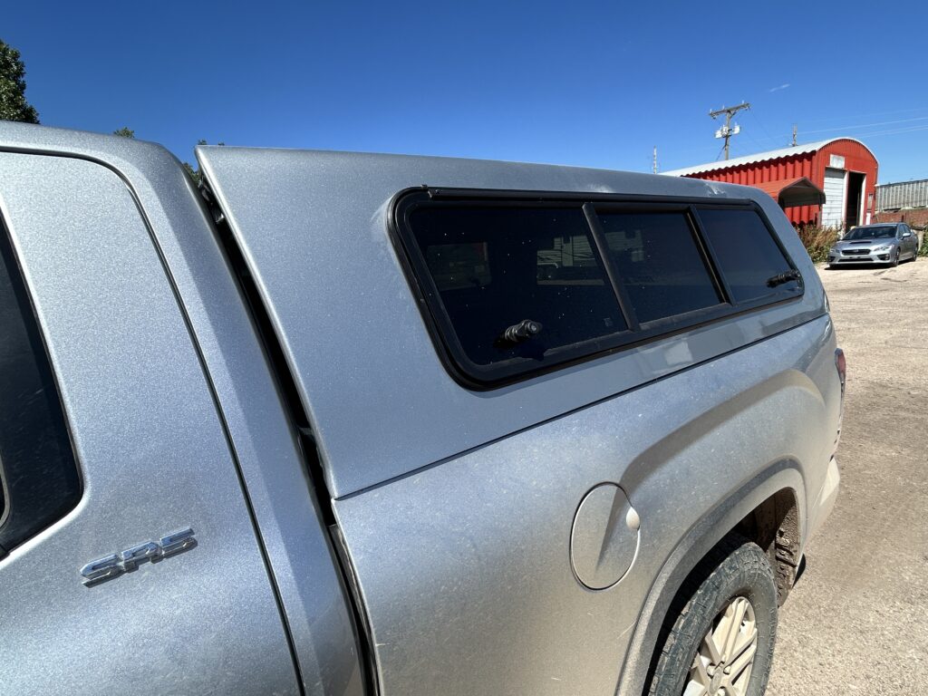 A silver Toyota Tacoma with an ARE truck topper installed, parked in a dirt lot near a red industrial building.