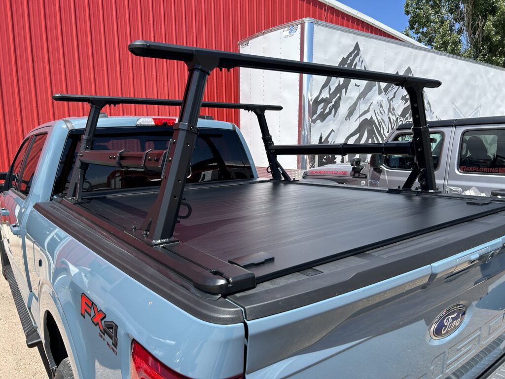 Adjustable black truck bed rack mounted on a blue Ford F-150 with a retractable tonneau cover, parked outside a red metal building.