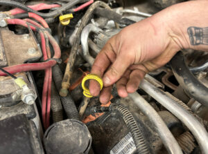 A mechanic's hand holding a broken and frayed engine oil dipstick tube in a dusty vehicle engine bay with red and black wiring and metal components.