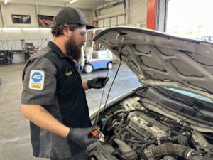 A bearded automotive technician wearing a black cap, black gloves, and an ASE Certified patch on his uniform checks the oil level of a car using a dipstick inside a garage.